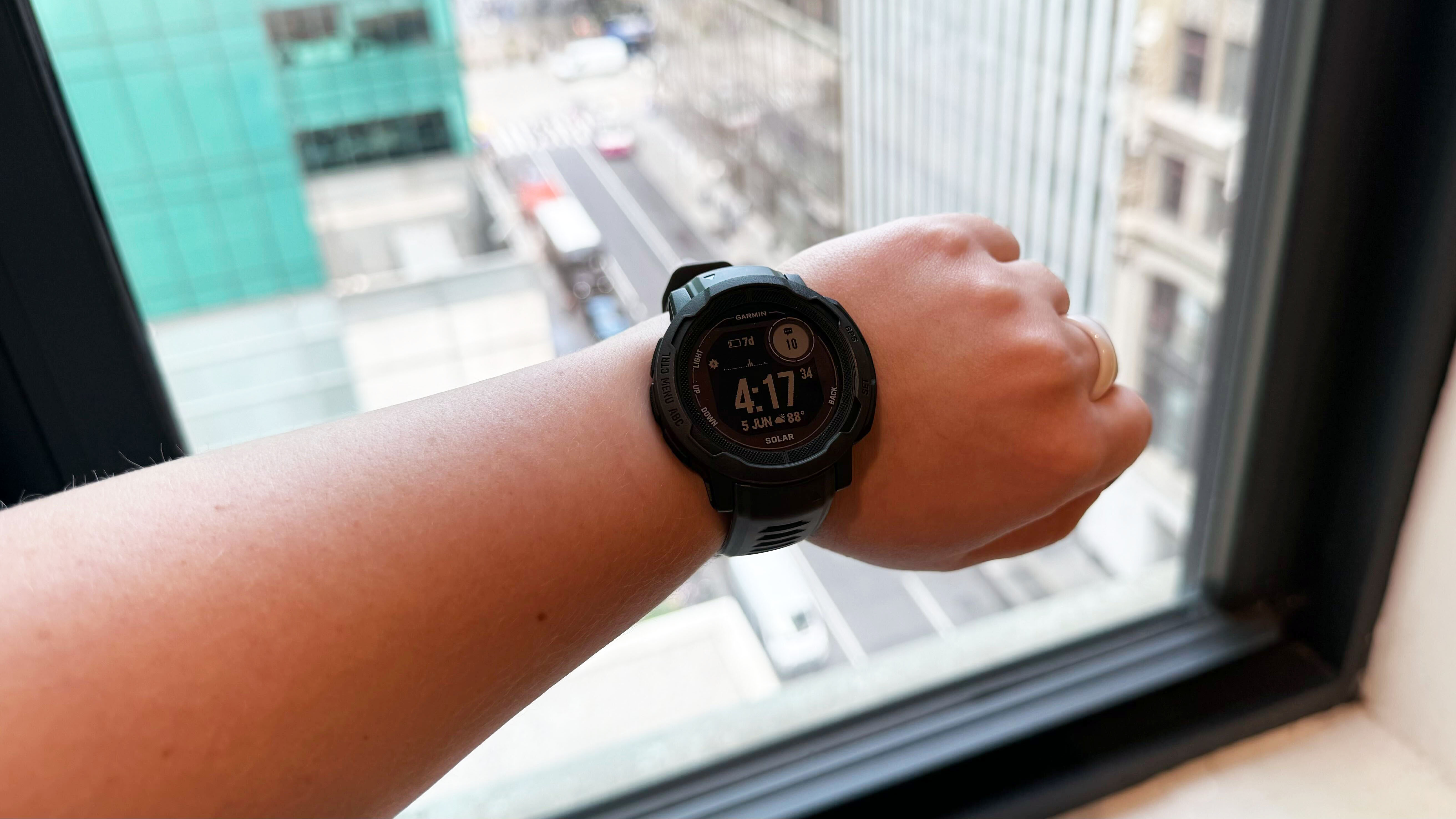 Garmin Instinct 2 Solar on left wrist, in front of a window overlooking Bryant Park in New York City.