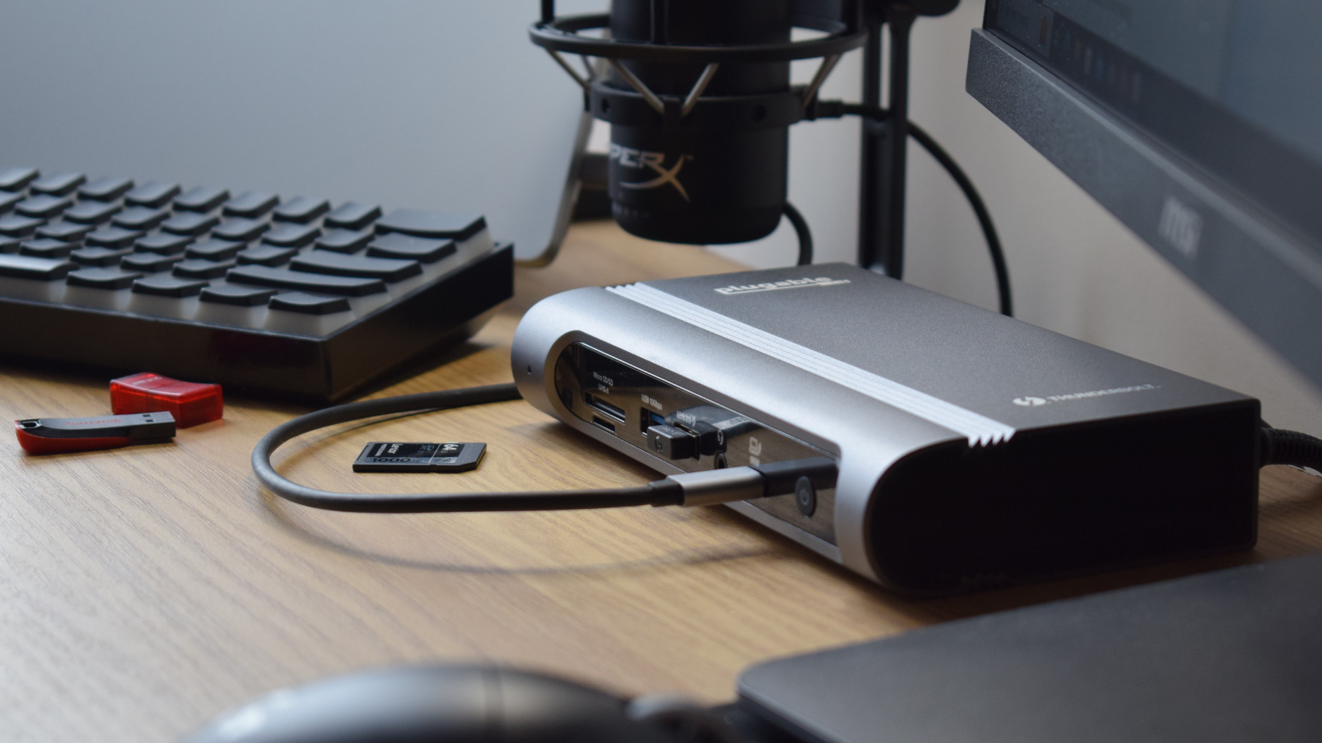 Photo of the Plugable Thunderbolt 4 & USB4 Quad Display Docking Station (TBT4-UDZ) as part of a desk setup connected to a laptop, microphone, external display, and wireless keyboard.