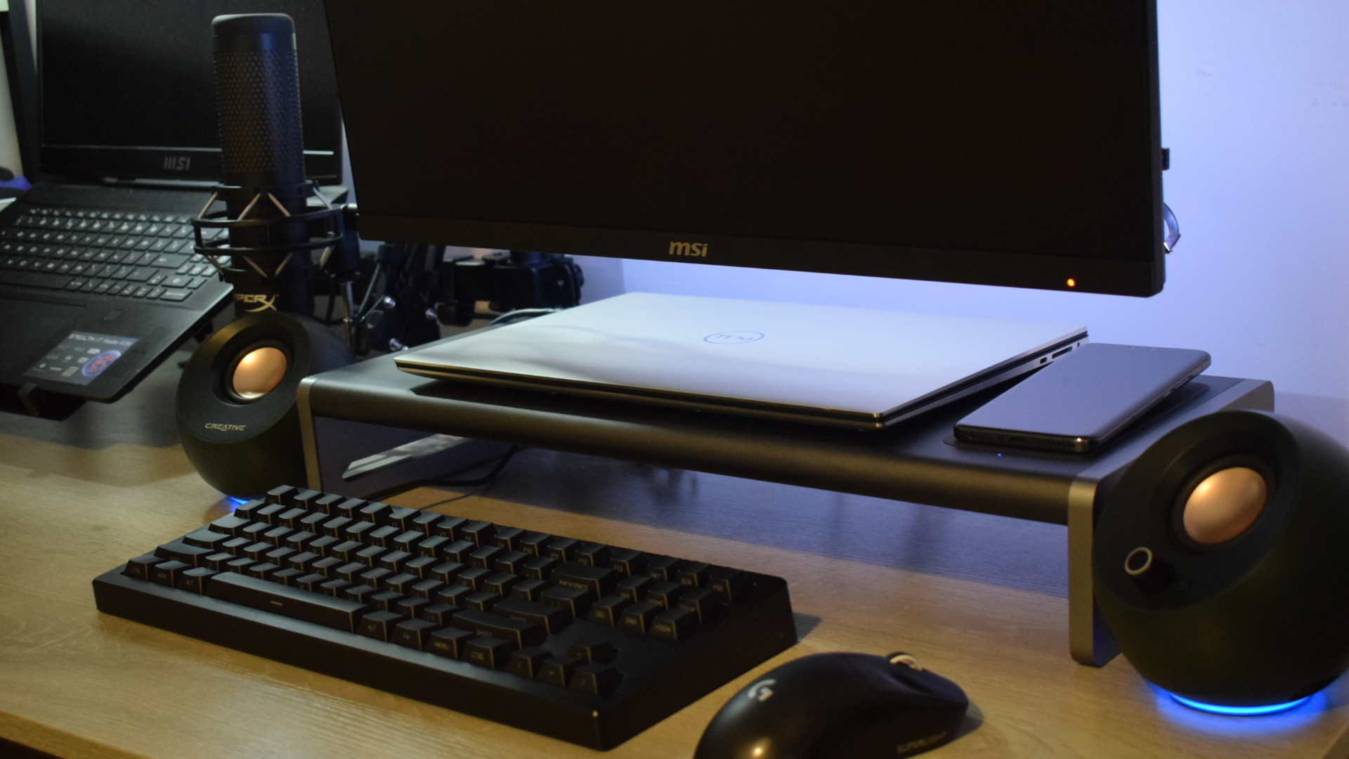 Photo of the Anker 675 USB-C Docking Station as part of a setup showing Creative Pebble speakers, a Microsoft Surface laptop, a Logitech wireless mouse and a wireless keyboard. An Honor Magic5 Pro smartphone is charging on the dock's wireless charging pad.