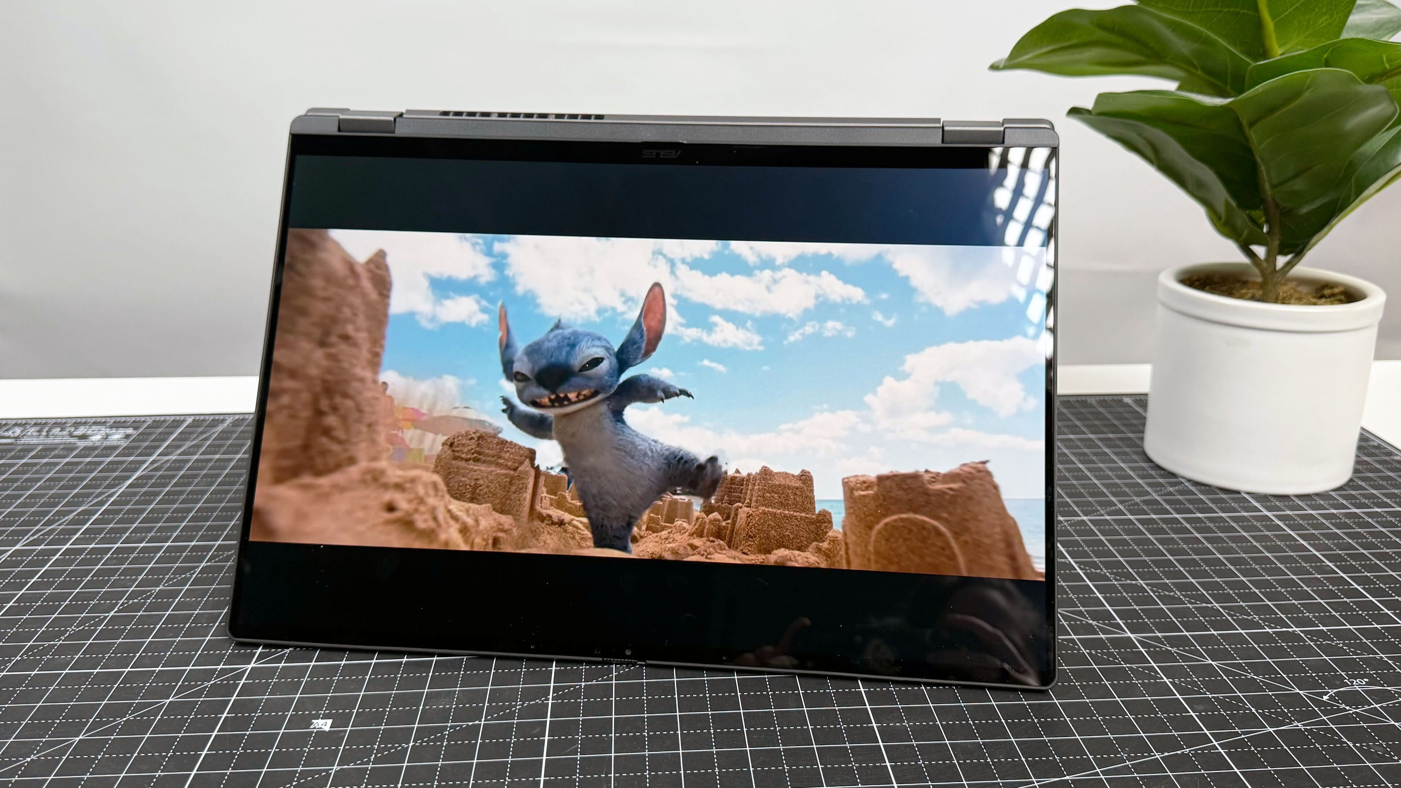 The Asus Vivobook 16 Flip convertible laptop in tent mode, playing a video. The laptop stands on a black and white desk against a gray background.