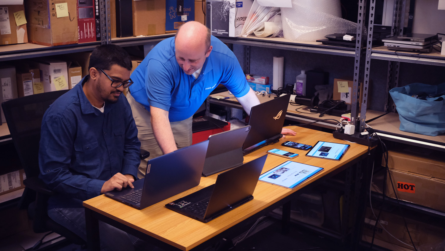 Two people in the Laptop Mag testing lab, with four laptops open in front of them on a wooden table.