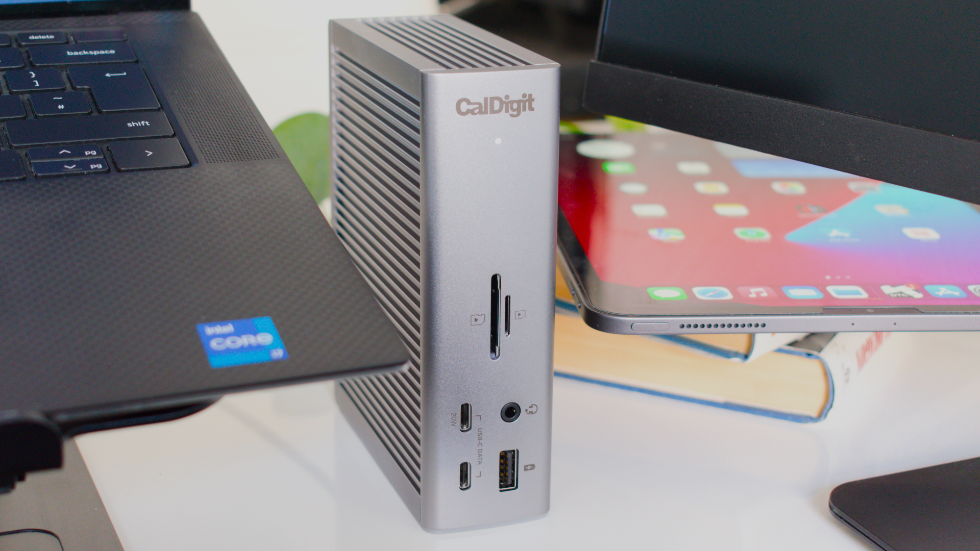 Photo of the CalDigit Thunderbolt Station 4 docking station showing the device on a white table next to a Dell XPS 15 laptop, an iPad Pro on top of a stack of books and an MSI monitor.